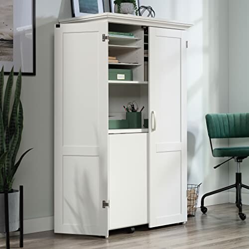 A white office storage cabinet with open doors, showing shelves and office supplies, beside a green chair.